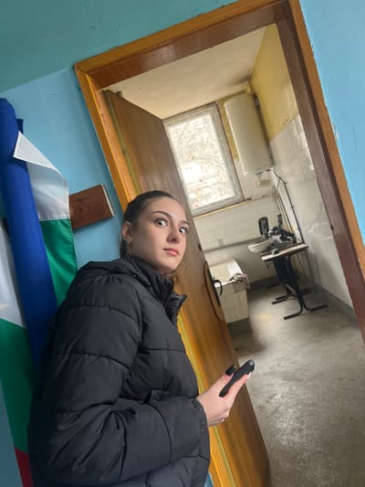 This image, taken in Cherven Bryag, Bulgaria, depicts a young female standing in a light blue hallway, looking into an adjacent utility room through a partially open wooden doorframe. The female, positioned to the left of the frame, has dark hair pulled back, wide eyes, and a slightly surprised expression. She wears a black puffer jacket and holds a black smartphone in her right hand, displaying manicured nails with black polish and light tips. To her immediate left, a Bulgarian flag (white, green, blue vertical stripes) hangs on the light blue wall, secured by a dark wooden plank.

The room visible through the doorway features yellow-painted walls above white tiling. A large window with a white plastic frame is visible on the far wall. A white cylindrical water heater is mounted on the yellow wall above the window. Below the window, a white utility sink or basin is partially visible. To the right of the sink, a dark metal stand supports a piece of industrial equipment, possibly a pressing machine or specialized apparatus, with visible wiring. The floor of this room is light gray concrete.