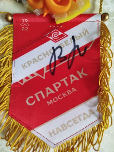 A red and white pennant with gold fringe hangs from a gold bead. The pennant features the text "КРАСНО-БЕЛЫЙ", "СПАРТАК", "МОСКВА", and "НАВСЕГДА". There is also a logo with a soccer ball inside a circle and a stylized infinity symbol with the numbers "19" and "22" above it. A black marker signature is present on the pennant. The pennant is laid out on a light-colored, textured surface, possibly fabric. The background is slightly blurred. There are no people visible in the image. The item appears to be decorative, possibly a souvenir or sports memorabilia. There is no information in the image that indicates its location in the city of Кузьмичи, Russia.