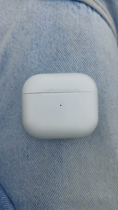 A white wireless earbud charging case is centered on a blue denim fabric. The case has a subtle seam running horizontally across its upper half and a small black dot, likely an indicator light, below the seam. The denim has a visible weave pattern and appears to be part of a garment such as jeans or a jacket. There are no people or other objects clearly visible in the image. The scene does not provide any information that would allow for the determination of its location in the city of Mersin, Turkey.