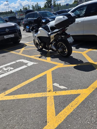 This media file shows a parking lot with several vehicles, including a white motorcycle with a black top case, a white car, and other dark-colored vehicles. The motorcycle is parked on asphalt marked with yellow lines, suggesting a designated parking area, possibly a no-parking zone or a specialized spot. There's a white wheelchair symbol painted on the asphalt, indicating a disabled parking space. The scene appears to be outdoors on a sunny day, with a clear blue sky and white clouds visible in the background. Trees line the edge of the parking lot.
The license plate on the motorcycle reads "7337 BE 150 RUS". Another license plate visible on one of the cars reads "X995XE 797". The image captures a static scene of parked vehicles, with no people actively engaged in any activity. The lighting suggests it is daytime, likely mid-morning to afternoon given the bright sunlight and distinct shadows cast by the vehicles. The overall impression is one of a typical parking lot setting on a pleasant day in Sholokhovo, Russia.