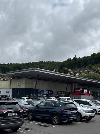 The image displays an outdoor scene of a commercial parking lot with a multi-story building alongside it, backed by a lush, forested hill under an overcast sky. Several cars are parked in designated spots.
In the foreground, a black Toyota RAV4 Hybrid with a Swiss license plate "AG-105178" is prominently visible. Next to it is a dark blue Skoda SUV with the Swiss plate "AG-127833 CH". Further down the row, a white Dacia Duster bears the German license plate "WTXT 669". Other vehicles are partially visible, including a white van and another SUV. One person can be seen near the black Toyota RAV4, appearing to be walking or standing.
The adjacent building features various storefronts and signs. Prominently displayed are signs for "WURST TEUFEL" (Sausage Devil) and "HÖLLISCH LECKER" (Hellishly Delicious), suggesting a food establishment. A large "K" logo is also visible, often associated with a German supermarket chain. Other visible texts include "Bären Apothek", "Apothek", "Raxon", "Audifahrt", "Mister Press", and "RUEDI TINNER AG BADEN".
The scene suggests a typical daytime shopping or commercial area, likely located near the German-Swiss border given the mix of German (WT for Waldshut-Tiengen) and Swiss (AG for Aargau) license plates, consistent with the provided context of Waldshut, Germany. The sky is completely covered with thick grey clouds, indicating an overcast or potentially rainy day.
