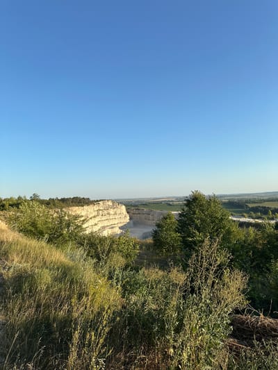 A high-angle, wide shot captures a bright, clear blue sky above a quarry and surrounding landscape. The quarry, a large, excavated area with layered, light-colored rock walls, is visible in the mid-ground. Green trees and overgrown vegetation occupy the foreground and frame the quarry on the left. The background shows rolling hills and fields stretching into the distance, with hints of buildings and structures suggesting a rural or semi-rural environment. The scene is illuminated by natural sunlight, casting shadows and highlighting the textures of the rock and foliage. The location is identified as a quarry in Verbka, Ukraine.