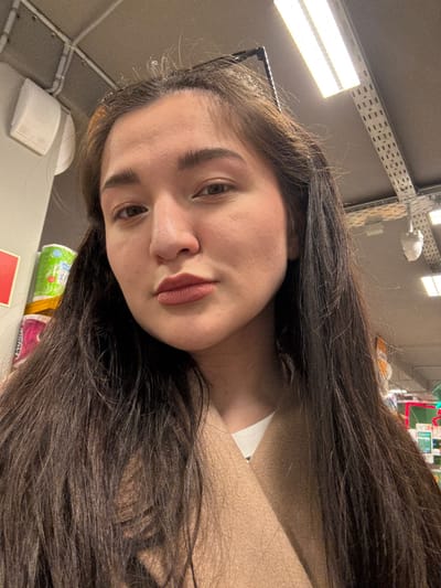 A young adult female is depicted in a close-up, chest-up shot within a retail store environment located in Kurovskoye, Russia. She has long, dark brown hair, parted slightly off-center, cascading over her shoulders. Her face is presented frontally, with light brown eyes gazing directly at the viewer. She exhibits a neutral expression with a slight pout, wearing a muted pink or nude lipstick. Her attire consists of a light beige or tan woolen-texture coat worn over a white, ribbed long-sleeved top, partially visible at the neckline.

The background, largely out of focus, reveals elements of a commercial interior. To the viewer's left, stacks of wrapped consumer goods, including items with green and pink packaging, are partially visible. A portion of a red sign or fixture is also present. To the viewer's right and above, the ceiling features exposed utilities: a network of silver-colored cable trays or conduits, rectangular fluorescent light fixtures providing artificial illumination, and a white, dome-shaped security camera or sensor mounted on the ceiling. The overall lighting is bright and even, consistent with an indoor retail space.