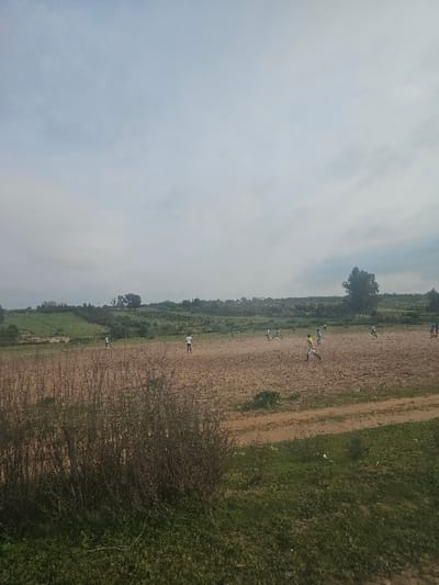 The image depicts an outdoor scene in Ameur عامر, Morocco, under an overcast sky with light grey cloud cover. Approximately 11-12 males are observed engaged in a game of soccer on an unpaved, reddish-brown dirt field.
The foreground consists of dense, dark green shrubs on the lower left, transitioning into shorter green grass and patches of bare earth on the lower right, where a dirt path or track is visible.
The midground is dominated by the expansive, dry, and uneven reddish-brown dirt field, which lacks formal pitch markings. Individuals, primarily wearing light-colored tops (white, yellow) and darker bottoms, are distributed across this field. One male in a yellow shirt and dark shorts is running forward in the mid-right, positioned near a light-colored football. Other players are scattered across the field, indicating active participation in the game.
The background features green, undulating terrain or agricultural land, sparsely populated with trees. Faint, indistinct structures are discernible on the distant horizon line to the far left.