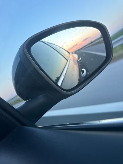 The image is a close-up shot of a car's side-view mirror. The mirror reflects a highway scene with a colorful sunset in the background. The sky is a gradient of blue, orange, and pink. The road appears to be a highway with guardrails visible on the right side of the reflection. The mirror itself has water spots on its surface. The interior of the car is visible in the foreground, showing a dark, textured surface, likely part of the door panel. The location of Palauzovo, Bulgaria is not depicted in this image, nor is any information provided that would place it there.