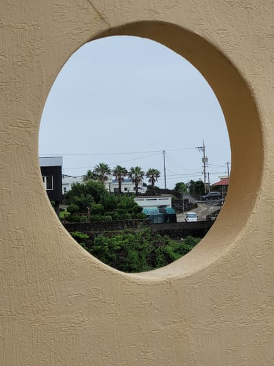 A wide-angle, eye-level shot shows a view of Seogwipo-si, South Korea, through a large, textured, light beige circular opening. The opening is in the foreground and frames the distant scene. The distant scene consists of a street with a few vehicles, houses, palm trees, and lush greenery. On the left side of the framed view is a modern, dark gray rectangular building with a white-framed window. Further into the distance, several white buildings are visible, some with red roofs. Four tall palm trees stand in a row between the dark building and the white structures. The street curves through the landscape, and several utility poles with a network of wires are prominent against the overcast sky. A dark-colored SUV is parked on the side of the road, and a white pickup truck is visible further down. The foreground wall, within which the circular opening is present, has a rough, stucco-like texture. The sky is a pale, uniform gray, suggesting an overcast day.