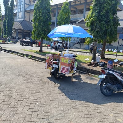 The image depicts an outdoor scene in Kendal, Indonesia. In the foreground, a paved walkway is visible, with a motorcycle and a food cart on it. The food cart, decorated with banners advertising "Telur Gulung" (rolled eggs), is mounted on a motorcycle. A blue umbrella is set up over the cart.
In the midground, a road runs alongside the walkway. Several people are seen walking and standing near a black pickup truck. Further back, a building complex with a distinct architectural style, likely a public facility or stadium, forms the backdrop. Tall trees line the area between the walkway and the building.
In the background, more people are visible, some sitting on steps and others walking. The environment appears to be a public space, possibly a park or an area adjacent to a large venue. The presence of the food cart and the casual arrangement of people suggest a lively, perhaps market-like, atmosphere.