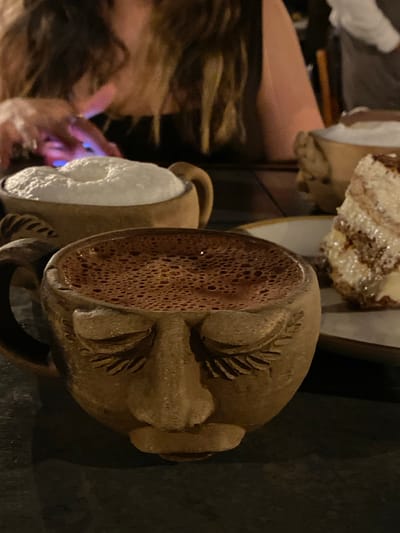 In Oaxaca City, Mexico, a blurred woman uses a phone at a table with dessert. Two textured, face-shaped mugs, one with white liquid and the other with brown liquid, are on the table. The setting is dimly lit.