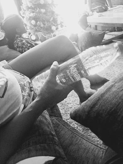 The monochrome image depicts an indoor scene in Ikate, Nigeria. In the immediate foreground, a person's right arm and hand are visible, holding a clear plastic bottle labeled "AQUAFINA." This person wears a light-colored short-sleeved top and blue denim jeans. A bare foot is partially visible on the carpeted floor. In the midground, another individual is reclining, with their bare legs prominently positioned. This person is dressed in patterned shorts and a top, with their head visible in profile, facing left towards a brightly lit area. Behind this individual, a decorated Christmas tree adorned with ornaments and lights is prominently displayed. In the background, elements of furniture and what appears to be another person's lower legs are faintly visible, illuminated by strong ambient light. The floor throughout the visible area is carpeted.