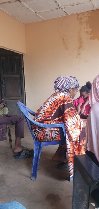 The image depicts an intimate interaction between an adult and a small child within an indoor or sheltered setting in Idah, Nigeria. An adult is seated on a blue plastic chair, wearing a vibrant orange, black, and white patterned garment, possibly a dress or wrapper, and a checkered headwrap. Their face is visible in profile, looking up at a young child in a pink top, who is leaning into or being held by the adult. The child's profile is also visible, looking down at the adult, suggesting a tender and affectionate moment.
The background features light brown or tan walls with noticeable areas of discoloration and water stains, indicative of an older building or maintenance issues. To the upper left, a white ceiling or panel with a grid-like pattern is visible. A dark, possibly metal, door or cabinet structure can be seen on the lower left. The floor is a light, neutral-toned concrete or dirt surface. Partially visible in the foreground are the legs and feet of another person, wearing dark pants and open-toed sandals, seated on a chair next to the main subjects. The lighting is diffused, suggesting a daytime scene. No visible text is present in the image. The scene conveys a sense of familial warmth in a modest local environment.