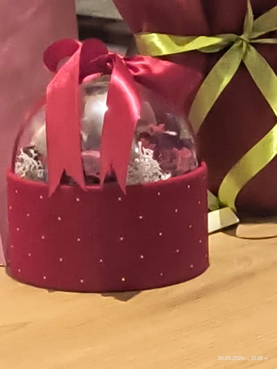 The image presents a still life composition featuring a maroon gift-wrapped box with a light green ribbon. Positioned beside it is a glass ornament encased in a maroon ribbon with small white dots. The ornament is adorned with a tied, bright red ribbon. The surface upon which these objects rest appears to be wooden. The date stamp "30.05.2026 r., 21:58 ч." is visible in the upper right corner of the image. The location of Dechkovtsi, Bulgaria, is not visually confirmed in the scene.