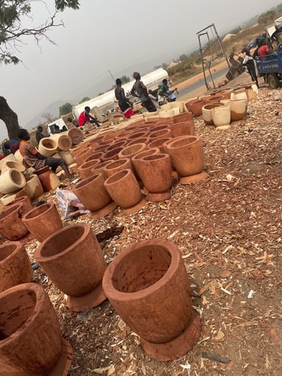 This image depicts an outdoor woodworking workshop or marketplace in Zongon Daji, Nigeria, under a hazy daytime sky. The ground is a reddish-brown earth heavily littered with wood shavings and sawdust. In the foreground and extending into the midground, numerous wooden mortars of varying sizes and finishes are arranged, predominantly cylindrical with some featuring a distinct base. These range in color from lighter natural wood to a reddish-brown.
Approximately 10-15 adult males are visible throughout the scene. In the left foreground, one man in a patterned orange and brown shirt is seated on a wooden object, handling another, apparently engaged in carving or finishing a mortar. Further back, several men are actively working on wooden items, suggesting the manufacturing or finishing of the mortars. One man in a red shirt is seated on a low wooden structure, working. Others are standing, observing, or moving within the busy workspace.
In the background, a paved road runs horizontally across the frame. A large white tanker truck, possibly labeled "AIR OIL," is parked or slowly moving on the road. To the right of the tanker, a smaller blue pickup-style truck is partially visible, with men handling wooden objects near its bed, possibly loading or unloading. Distant hills are faintly visible against the hazy horizon. A large tree with sparse branches frames the upper left portion of the scene.