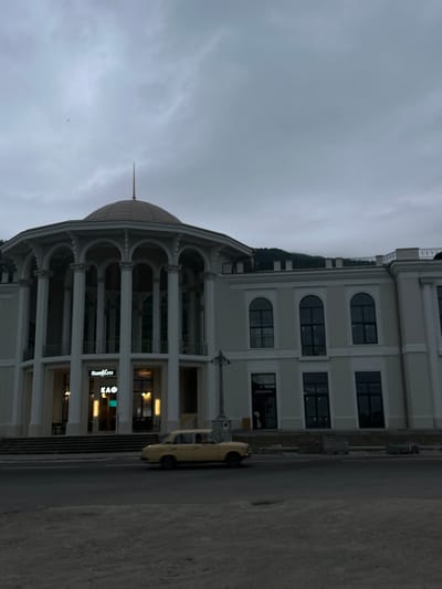 The image depicts the "Sanli.co" cafe and the adjacent building in New Athos, Abkhazia, under a cloudy sky. In the foreground, a yellow, older model sedan is driving on the asphalt road. The building features a large, domed portico with multiple columns and arched windows. The cafe's signage, "Sanli.co" and "КАФ," is visible. Behind the cafe, part of a larger building with arched windows is visible, extending to the right. The background shows a dark, overcast sky and the silhouette of a mountain range. The overall scene conveys a somber and somewhat retro atmosphere.