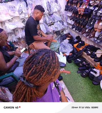 This image captures an indoor scene within a shoe store or market stall in Agbani, Nigeria, featuring three individuals. In the center, a man with short, styled hair sits on a low stool, wearing a black t-shirt and brown shorts. He is intently focused on an orange shoe or sandal he holds in his hands, seemingly engaged in repair, cleaning, or preparation.
To the left, a woman is partially visible, seen from behind and the side. She wears a leopard-print headscarf or bandana and a dark top, looking down at a mobile phone in her hands. In the immediate foreground, with her back to the viewer, is another woman with dark and light brown twisted braids, pulled back, and wearing a purple top. She appears to be looking towards the right side of the frame.
The setting is densely packed with footwear. The walls are lined with numerous shoes, mostly women's, individually packaged in clear plastic bags. Many of these bags have "TOP" printed on them. On the right, a wide variety of sandals, slides, and casual shoes are arranged on shelves and directly on the floor, some presented in small cardboard boxes, including distinctive orange ones. The floor in the foreground is covered with green artificial turf. The environment appears brightly lit, likely by artificial light.
Visible text includes "TOP" on the plastic shoe bags, "CAMON 40" as a camera watermark on the bottom left, and camera exposure information "23mm f/1.8 1/50s ISO892" on the bottom right.
