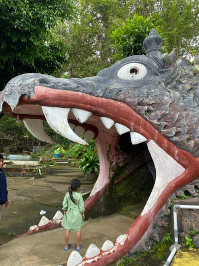 A large, open-mouthed, concrete sculpture of a dragon-like creature is located in the city of Kecamatan Manyaran, Indonesia. The sculpture has a black and grey body with red and white details, and a bright white mouth filled with white teeth. The mouth is open, forming a large, dark opening leading into the sculpture. A young girl with long dark hair, wearing a green dress, stands in front of the sculpture, facing the camera. An adult male in a blue shirt stands behind her, looking away from the camera. The ground around the sculpture is made of concrete and covered in green moss. Lush green vegetation surrounds the sculpture. The scene is located in the city of Kecamatan Manyaran, Indonesia, at the coordinates -7.826 latitude and 110.816 longitude.