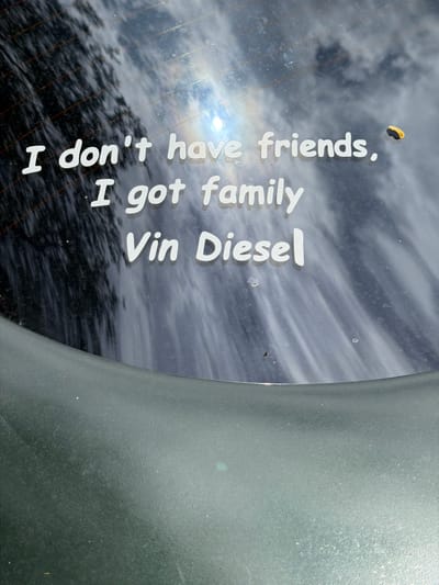 The image shows white text applied to the rear window of a vehicle. The text reads "I don't have friends, I got family Vin Diesel". The background appears to be a car wash with water streaks and reflections from the sun. A small yellow and black object, possibly a sticker or debris, is visible on the right side of the window, next to the word "friends". The bottom of the image shows a dark, textured surface which is likely the car's rear bumper or a part of the vehicle's interior. The context provided indicates that this scene is located in Reckange-sur-Mess, Luxembourg, though there are no visual cues within the image to confirm this specific location.