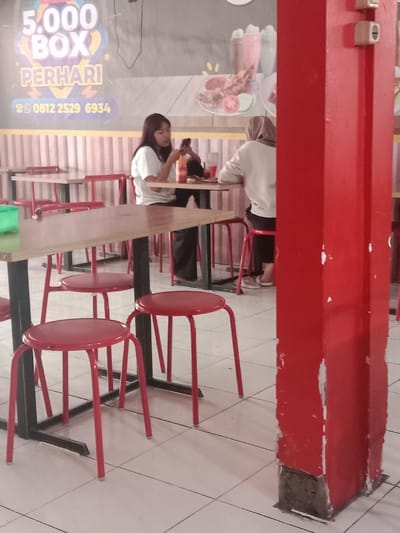 A brightly lit indoor dining area with white tile flooring. Two women sit at a table in the background, one looking at her phone. In the foreground, there's a table with red stools. A large, red, square support column dominates the right side of the frame, showing signs of wear and exposed base. A wall mural is visible behind the diners.