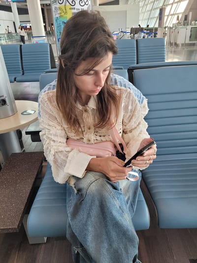 A woman sits in an airport waiting area, looking at her phone. She has long brown hair and is wearing a white lace blouse and ripped jeans. A pink purse is slung across her chest. The background shows rows of blue airport seating and a large window overlooking the city. The scene appears to be at Incheon International Airport, as indicated by Korean signage in the background and the architectural style typical of major international airports.
