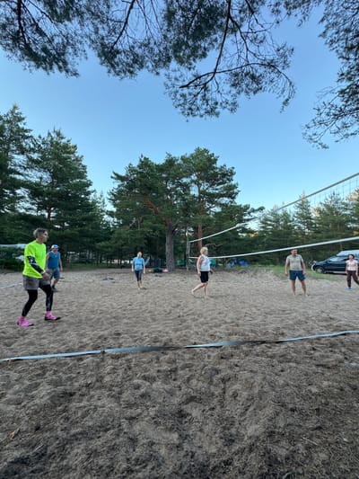 In Ozerki (Krasnodolinskaya), Russia, a group of adults is playing volleyball on a sandy court surrounded by pine trees. The sky is clear and blue, indicating it is daytime. The individuals, some dressed in athletic wear, are positioned on both sides of the volleyball net, actively engaged in the game. In the background, several tents and a car are visible, suggesting a recreational area or campsite. The sand on the court shows footprints, indicating recent activity. The overall scene depicts a casual outdoor sporting event.