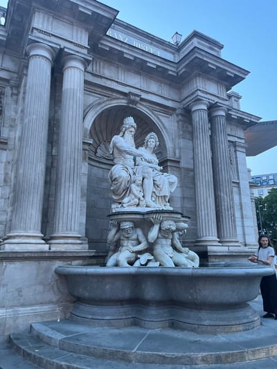 The image shows a fountain in front of a building in Vienna, Austria. The building is made of stone and has a large archway. The fountain is decorated with a group of statues, including two figures sitting on a pedestal and two mermen at the base of the fountain.  The figures are made of white marble and depict a man with a beard, a woman with a crown, and a child. The inscription "FRANZ JOSEF I" can be seen above the archway. A woman in a black shirt, gray pants, and a black backpack is standing to the right of the fountain. She is looking at the fountain. The sky is blue and the sun is shining. The fountain is located in front of the Museum of Art History, which is located at the coordinates 48.204, 16.369.