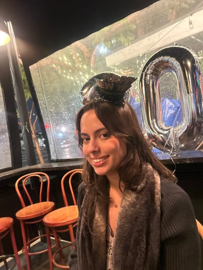 An image taken in Culver City, United States, depicts a single female subject in the foreground, smiling directly at the camera. She has long, dark brown hair and wears a small black "Happy New Year" party hat headband. Her attire includes a dark ribbed sweater, a light-colored, possibly sequined top, and a brown faux fur scarf or collar. A delicate necklace with a small pendant is visible.

Behind the subject, to her right, a large silver foil balloon shaped like the number "0" is prominently displayed, suggesting a celebration. To her left, at least three wooden bar stools with round, light-colored seats and reddish-brown frames are visible, consistent with a casual dining or bar setting. A dark countertop or bar surface spans the mid-ground.

Beyond the countertop, large windows or a semi-enclosed outdoor patio area are visible, covered with transparent plastic sheeting that shows streaks of condensation or moisture. Through this transparent barrier, a blurred nighttime urban street scene is observed. Trees adorned with string lights are visible, along with distant, blurred vehicle headlights and streetlights. The environment is an indoor commercial establishment, likely a restaurant or bar, with a festive, nocturnal ambiance.