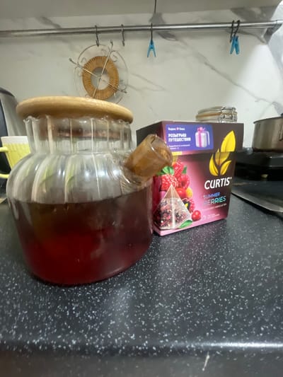 A close-up, slightly blurry, rotated shot shows a tea pitcher filled with a dark red liquid, likely tea, next to a box of "CURTIS SUMMER BERRIES" flavored hibiscus tea. The tea box has an illustration of raspberries and other berries, and text indicating it contains 20 tea bags. Part of a label on the box reads "Яндекс + Плюс РОЗЫГРЫШ ПУТЕШЕСТВИЯ," suggesting a promotional giveaway. The scene appears to be indoors, possibly a kitchen, with a speckled countertop and a light-colored, possibly marble, backsplash. Two blue clothespins are visible hanging on a metal rack in the background. The lighting suggests it could be daytime.