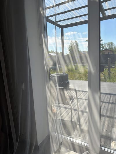 The image depicts an outdoor patio area with a wooden deck, viewed through a glass door and sheer curtains. Sunlight streams through a translucent roof structure, casting diagonal shadows across the scene. On the deck, a metal chair and a stack of tires are visible. In the background, there is a grassy area with some trees and a building. The overall impression is one of a residential backyard on a sunny day. The location is identified as Malinovka, Russia.
