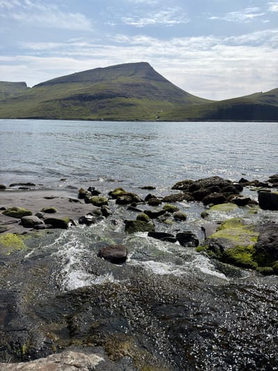 The image depicts a rocky shoreline with calm water in the foreground, leading to a body of water with gentle waves. In the background, rolling green hills rise, with a prominent, pyramid-shaped mountain dominating the landscape. The sky is partly cloudy, with streaks of white clouds against a pale blue. The shoreline is characterized by dark, wet rocks, some covered in vibrant green moss, interspersed with black sand. Sunlight reflects off the water, creating a shimmering effect. The scene appears tranquil and natural, possibly representing a view from the village of Bøur in the Faroe Islands, known for its dramatic coastal scenery and mountainous terrain. There are no people or animals visible in the image, nor any specific man-made structures that are clearly identifiable. The overall impression is one of natural beauty and serene isolation.