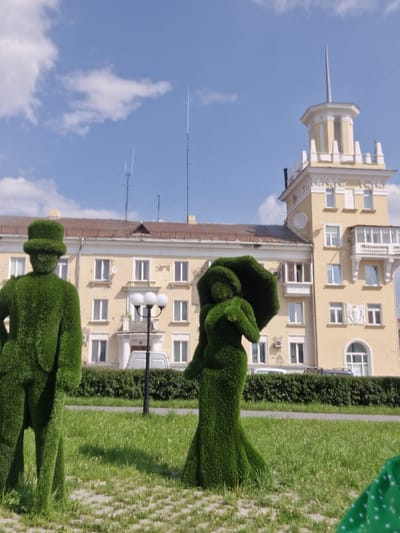 The image shows a sunny day in Miass, Russia, with a pale yellow building in the background that features a tower with a spire. In the foreground, two topiaries, one resembling a man in a top hat and the other a woman holding an umbrella, are situated on a lawn in front of a paved area. A lamppost with three globe lights stands between the topiaries and the building. A white van is parked in front of the building, and a dense green hedge runs along the perimeter of the lawn. The sky is blue with scattered white clouds.