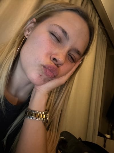 A blonde woman with freckles poses closely for a selfie. She has her eyes closed, puckers her lips, and rests her face against her hand. A gold watch is visible on her wrist and a large gold hoop earring is in her ear. She wears a black shirt, and light from above illuminates her face. White curtains and other household objects are visible in the background.