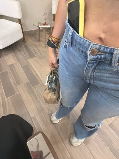A woman sits indoors on a tiled floor, holding a Starbucks bag. She wears light-wash, ripped jeans and white sneakers. A yellow smartphone protrudes from her open jeans. Her midriff is exposed. She has bracelets on her left wrist. In the background are two white chairs, a small table, and a partial view of a vanity. The scene is in Villahermosa, Mexico.