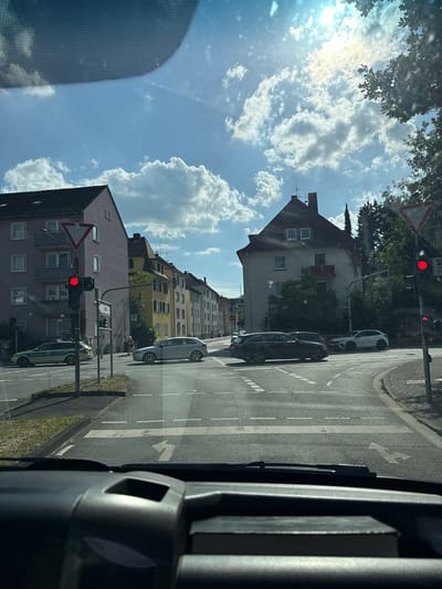 The image depicts a view from inside a vehicle, looking out at a street scene in Giessen, Germany. The perspective is slightly tilted, suggesting the vehicle is moving or parked on an incline. The sky is a bright blue with scattered white cumulus clouds, indicating it is daytime and the weather is fair. Sunlight illuminates the scene, casting shadows and creating highlights.

The street is paved and marked with white lane lines, including a directional arrow. Several cars are visible on the road, including a white car, a dark colored car, and a silver car. A police car with markings is also present. There are buildings lining the street, some appearing residential with multiple windows and balconies. Trees with green foliage are interspersed along the street and near the buildings.

A traffic light is visible, showing a red signal, which is likely the reason for the vehicles being stopped. A red triangular warning sign is also posted near the traffic light. The interior of the vehicle is partially visible on the right side of the image, with a dark dashboard and what appears to be a portion of a steering wheel or console.

The overall impression is a typical urban street scene in Germany during the day, with traffic, buildings, and pedestrian infrastructure. There are no people clearly visible in the image.