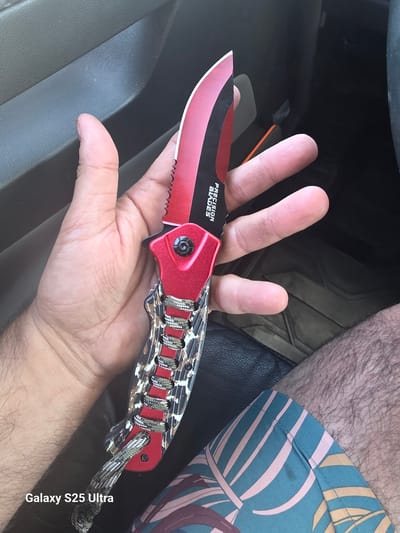 A close-up, eye-level shot shows a person's right hand holding a red and black folding knife. The knife has a jagged edge on the blade, and the handle has a camouflage pattern with gray cord lacing. The person's arm and leg are visible, and they are wearing patterned shorts. The background appears to be the interior of a vehicle, with a dark gray textured surface on the left and a black textured surface on the right. A watermark in the bottom left corner reads "Galaxy S25 Ultra". The image was taken in Spokane, Washington, United States.