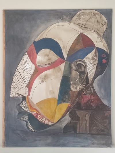 The image is a painting of a stylized human head in profile, facing left. The head is segmented into geometric shapes and filled with various colors and patterns, including blue, red, brown, yellow, and white with dot patterns. The background is a mottled gray. The subject's hair is in a bun on top of the head. The painting also features an ear with hanging earrings and some indistinct shapes behind the head and neck. The setting cannot be determined from the image and there is no evidence to suggest it is located in Muang Pattaya, Thailand.