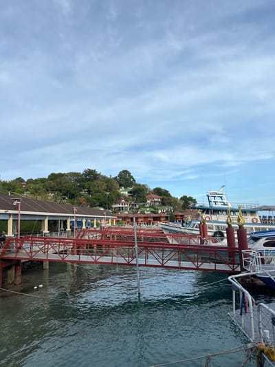 This is a photograph of a pier in Ban Haad Rin, Thailand. In the foreground, a red metal bridge spans the water, connecting the land to the boats docked at the pier. The water is a deep blue-green and appears calm, with ripples reflecting the sky. Several boats, including passenger ferries and smaller vessels, are moored alongside the pier. Some of these boats are white with blue accents and have life preservers visible. Behind the pier, a hillside is covered in lush green trees and dotted with colorful houses. The sky above is a light blue with scattered white clouds. The architecture of the pier structures includes dark-tiled roofs supported by columns. There are no people clearly visible in the image.