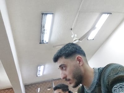 The image captures an indoor scene from a low-angle perspective, showing two men and parts of a room's interior. The original image is oriented sideways, with the subjects appearing upside down; however, the description will refer to an upright orientation.

In the foreground, a man is prominently featured in profile, looking towards the right side of the frame. He has short, dark, neatly styled hair with faded sides, and a well-groomed beard. He is wearing a thick, patterned sweater in shades of gray and green. His expression is neutral as he gazes slightly upwards and to his right. In the upper left corner, partially visible above the first man's head, is the top of a second man's head and shoulder, also wearing a similar patterned garment.

The background primarily consists of plain, light-colored walls or a ceiling. Several artificial light sources are visible: a white ceiling fan with multiple blades hangs towards the lower-middle section of the frame, and three long, rectangular fluorescent light fixtures are mounted on the wall. Two of these fixtures are clearly visible on the right side, emitting bright light, while a third is partially seen on the left near the ceiling fan. A wide, light-colored column or structural element runs vertically along the far right edge of the image. A grid-like ceiling structure is faintly discernible in the top right corner.

No specific activity or event is clearly taking place; the men appear to be casually looking or observing. The bright artificial lighting indicates an indoor setting, likely a public or semi-public room such as an office, classroom, or hall within a building. There are no visible texts or distinctive markings that would further specify the location beyond the given context of Ash-Shaghour Municipality, Syria. The overall atmosphere is neutral.