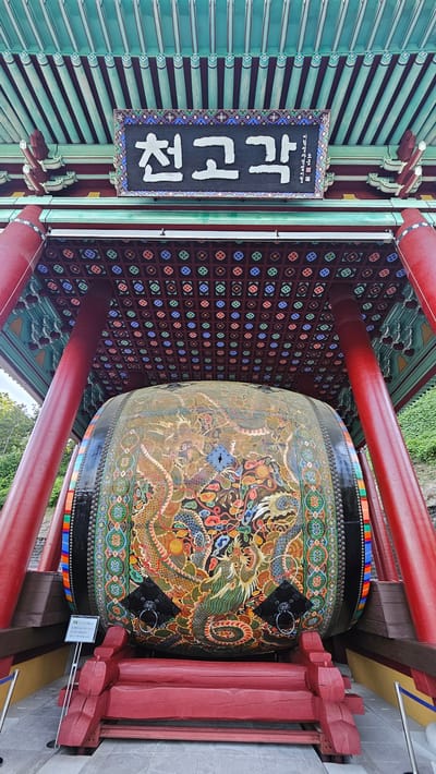 A low-angle, full shot captures a large, elaborately decorated drum situated outdoors under a traditional Korean pavilion structure. The pavilion features a teal-colored, tiered roof with dark brown eaves and red beams supporting it. The ceiling of the pavilion is adorned with a repeating pattern of small, colorful floral motifs in red, blue, green, and yellow against a dark brown background. A black sign with white Korean characters, "천고각", is prominently displayed above the drum. The drum itself is immense, painted with vibrant depictions of dragons, clouds, and intricate patterns in shades of green, gold, red, blue, and orange. Two black ring handles are attached to the sides of the drum. The drum is supported by a red wooden base. To the left of the drum, a white sign with Korean text and a blue rope barrier are visible. The background shows a green, grassy hillside.