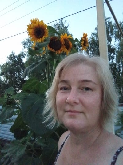 In Ворошиловка, Kazakhstan, a close-up shot captures a fair-skinned woman with blonde hair standing outdoors near several sunflowers. The woman is positioned in the foreground, facing the camera, with her face and upper chest visible. She has fair skin, light blue eyes, and her blonde hair is styled in a choppy, shoulder-length cut. She is wearing a dark top with a white floral pattern.
Behind her, a stalk of sunflowers with large green leaves is prominent. Several sunflower heads are visible, with yellow petals and dark brown centers. The background consists of trees and a pale blue sky with visible power lines. A light-colored pole is also present to the right of the woman. The overall lighting suggests it is either early morning or late evening.