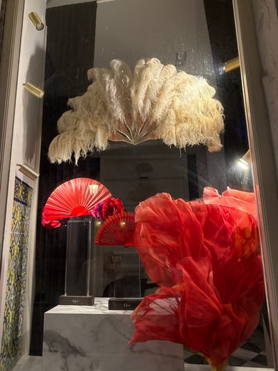 This media file captures a visually striking display, likely a shop window or exhibition, featuring two large, elaborate fans and a flowing piece of fabric. On the left, a fan made of numerous cream-colored feathers is displayed, its shape reminiscent of a large plume or a peacock's tail. The feathers are arranged in a semi-circular fashion, showcasing their delicate texture and volume. To the right, a voluminous piece of vibrant red or coral fabric drapes dramatically, appearing to be made of a sheer, lightweight material that catches the light and creates soft folds and shadows.
Beneath the large fabric, two smaller, traditional Spanish fans are visible. One is a deep red, partially open, adorned with what appears to be a large, dark pink or fuchsia rose at its center, with smaller red roses placed strategically. Another red fan is positioned slightly above and to the right, also partially open, suggesting a theme of flamenco or Spanish artistry.
The scene is illuminated by spotlights, which create bright highlights and deepen the shadows, emphasizing the textures and forms of the displayed items. The background is a mix of dark and reflective surfaces, with hints of what could be a marble-patterned display stand on the far right. The overall aesthetic suggests a theatrical or celebratory presentation, possibly related to fashion, dance, or cultural heritage.
The image gives a sense of opulence and drama. The setting appears to be indoors, given the artificial lighting. There is a tiled floor in the foreground with a yellow and blue pattern, hinting at a specific regional or cultural context. While no people are visible, the objects themselves evoke a sense of performance and allure. There is no visible text in the image. The composition and vibrant colors suggest this display is designed to attract attention and evoke emotion, perhaps wonder or excitement.