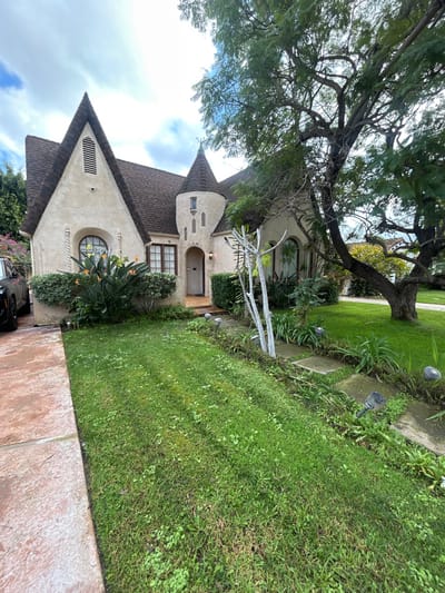 A fairytale-style house with a conical tower sits beneath a large tree. A dark roof, beige walls, and arched windows lend character. Lush greenery surrounds the house, with flowers blooming near the base. A green lawn stretches beside the house. Paving tiles are in the foreground. The sky is partly cloudy.