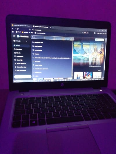 The image shows a laptop displaying a movie streaming website, "MovieBox". The search bar contains the letters "HI" and search suggestions like "Heartbreak High," "High Potential," "Hijack," are displayed. The laptop's keyboard and trackpad are visible. The lighting in the room is a purple hue, possibly from ambient lighting. The city of Juja ward, Kenya is not relevant, and does not appear to be contained in this scene.