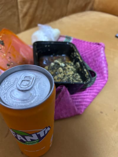 The image displays a close-up, elevated view of food and beverage items situated on a light brown, possibly wooden or upholstered, surface in Ilishan, Nigeria. In the immediate foreground, an upright, orange Fanta brand aluminum can is visible, covered in condensation. Its top, including the unopened pull-tab, is clearly detailed. Behind the Fanta can, towards the right, a black rectangular plastic food container holds partially consumed food consisting of a dark, viscous sauce or stew with lighter, granular solid components mixed within. This container rests upon a fuchsia-pink woven mat with a distinct striped pattern. To the left of the black container, an orange or reddish plastic item, possibly another container or bag, is present. Further back and slightly left of the black container, a small, clear plastic bag contains a white, amorphous substance. The background is out of focus. No individuals are depicted in the scene.