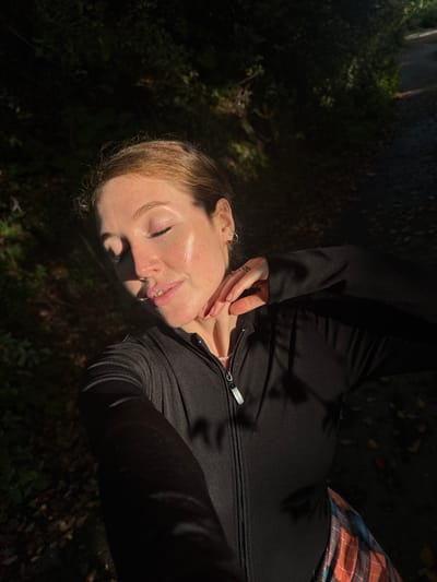 A woman is lying on the ground outside. She is wearing a black long-sleeve zip-up jacket and a brown plaid skirt. Her hand is raised towards her face. The surrounding ground is covered in leaves and shadowed by trees. The location is presumably a natural area but not identifiable within Tsanbaikwara, Abkhazia, from the visual information.