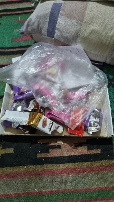 The image displays an indoor scene in Yangi-Khayat, Uzbekistan, depicting a collection of confectionery items within a shallow cardboard box. The box contains several open and crumpled candy wrappers, including a white and gold "Miller Capricho" wafer bar wrapper, purple wrappers with "JUST" visible on one, and a crumpled silver foil wrapper. A clear plastic bag filled with numerous individually wrapped candies, predominantly pink and red, occupies a significant portion of the box. A white paper or label with dark text is visible through the plastic bag. The box rests on a patterned rug or carpet featuring horizontal stripes and geometric designs in green, black, brown, red, and beige tones. A light-colored pillow with blue and beige stripes is partially visible in the upper left background. Small crumbs or debris are scattered on the rug in the foreground. No people are visible.