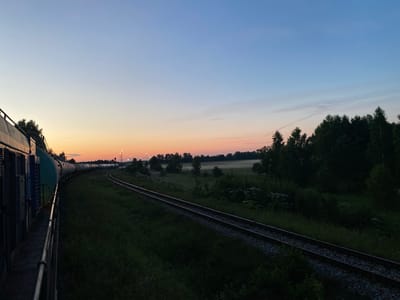 The image shows a train moving along a railway track during dusk. The sky is a gradient of pale blue to orange. On the left, the side of the train, consisting of multiple cars, is visible. The train is on a curved track that leads into the distance. The surrounding environment is a mix of grassy fields and trees. In the distance, there are some lights that suggest a town or industrial area. The overall scene conveys a sense of travel and transition during twilight. The location is Rychino, Russia.