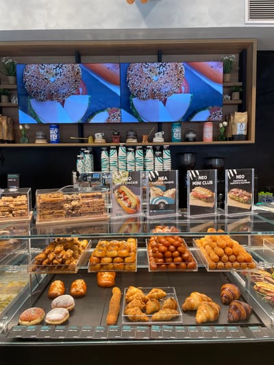 A display of baked goods is presented in a bakery. The baked goods include various types of pastries, donuts, and croissants, arranged in clear plastic containers and on a black counter. Behind the display, there are two large screens showing images of bagels. Various coffee cups, cans, and coffee machines are also visible on shelves above the display. The bakery appears to be located in Thessaloniki Municipal Unit, Greece, as indicated by the Greek text visible on some of the product labels and signage. The overall environment is clean and well-lit, suggesting a modern and professional establishment. There are no people visible in the image.