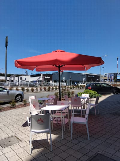 The image shows an outdoor seating area with tables and chairs in front of a row of businesses, under a large red umbrella. In the foreground, several white plastic chairs and tables are set up on a tiled patio. One chair, in the immediate foreground, has the "Brunati" logo in red on its back. In the background, several cars are parked and driving on a road. The buildings behind the road appear to be car dealerships, with visible signage for "PEUGEOT", "HYUNDAI", and "KIA". The sky is clear blue. This scene is located in Vila Nova de Famalicão, Portugal, specifically at the business park of Famalicão, on the road leading to the city center.