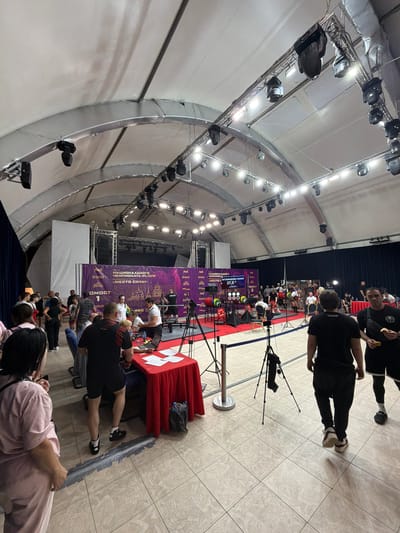 The image captures an indoor sporting event, likely a powerlifting competition, taking place within a large, tent-like structure. In the background, a stage is set up with a large purple banner displaying Cyrillic text, including "НАЦИОНАЛЬНОГО ЧЕМПИОНАТА IPL/СПР" (National Championship IPL/SPR) and "МЕСТО СИЛЫ" (Place of Strength). The stage is illuminated by numerous spotlights.
In the foreground and midground, a crowd of people are gathered. Some are spectators, while others appear to be athletes or event staff. A red-draped table is set up with papers and a laptop, suggesting an information or registration desk. Several tripods with cameras are positioned around the area, indicating filming or photography of the event. The floor is tiled, and a blue rope barrier separates different sections of the venue.
In the mid-left of the image, weightlifting equipment, including colored weight plates, is visible on a red carpeted platform. People are actively engaged in the competition or in discussions related to it. The overall atmosphere suggests an organized sporting event. The event is located in Suzdal, Russia.