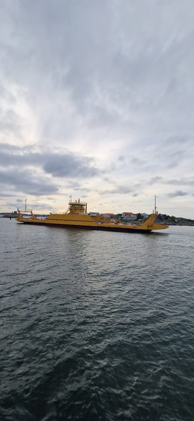 A yellow car ferry, the "GOTA", is viewed from the water on a cloudy day. The ferry is positioned horizontally across the frame, with its bow on the left and stern on the right. A multi-level superstructure with a bridge is visible in the center of the ferry. Several cars are parked on the deck of the ferry. In the background, a coastline with houses and trees is visible. The water is choppy with visible ripples and waves. The sky is overcast with gray clouds. The image appears to have been taken from a boat or the shore. The location is likely Öckerö, Sweden, based on the ferry's name, which is a common name for ferries operating in the Gothenburg archipelago.
