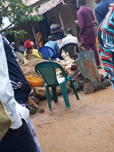 In Panyam, Nigeria, a group of people are gathered outdoors. A corrugated metal roof shelters some individuals from the elements, while others stand and sit in the open. Among the seated are individuals in blue and black plastic chairs. The ground is covered with sand and scattered debris, including a broom and rocks. A large, rusted metal drum stands prominently in the scene. Clothing worn by the individuals varies, with one person in a burgundy outfit, another in a blue and white striped shirt, and a woman with a red headscarf. The overall impression is of an informal gathering or event in a semi-urban or rural setting.